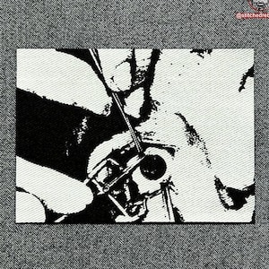 May include: Black and white image of a hand holding a needle and thread, sewing a piece of fabric. The fabric is textured and has a design on it.