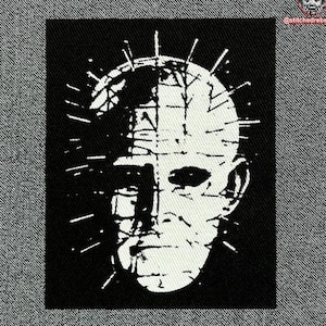 Op de afbeelding: Zwart-witte geborduurde patch van een gezicht met een kroon van doornen. Het gezicht is gestileerd en heeft een scherpe, hoekige uitstraling.