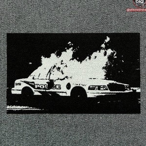 May include: Black and white image of a police car engulfed in flames. The car is a white saloon with the word "POLICE" written on the side. The flames are large and engulf the entire car.