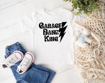 May include: White t-shirt with the text "Garage Band King" in black, accompanied by a black lightning bolt graphic. The shirt is paired with blue jeans and white sneakers with red accents. A stuffed bunny and greenery are also in the image.