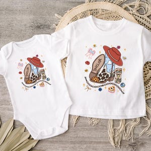 Op de afbeelding: Een witte romper met korte mouwen en een wit T-shirt met korte mouwen, beide voorzien van een grafische print van een cowboylaars met een rode hoed, de naam Taylor, een blauwe luchtpatroon, koeienprint en kleine decoratieve iconen.