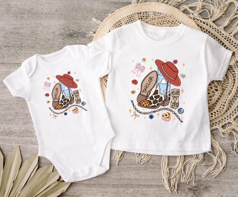Op de afbeelding: Een witte romper met korte mouwen en een wit T-shirt met korte mouwen, beide voorzien van een grafische print van een cowboylaars met een rode hoed, de naam Taylor, een blauwe luchtpatroon, koeienprint en kleine decoratieve iconen.