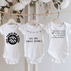 May include: Three white baby onesies hanging on wooden hangers. Each onesie has a different black graphic and text design. One says "All Panic No Disco", another says "All The Small Things", and the last says "Do you have the time to listen to me whine..."