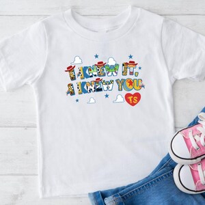Puede incluir: Una camiseta blanca de manga corta con el texto I KNEW IT, I KNEW YOU en letras de bloque coloridas y estampadas. El dise&ntilde;o incluye peque&ntilde;os sombreros de vaquero rojos, estrellas azules, nubes blancas y un coraz&oacute;n rojo con las letras TS.