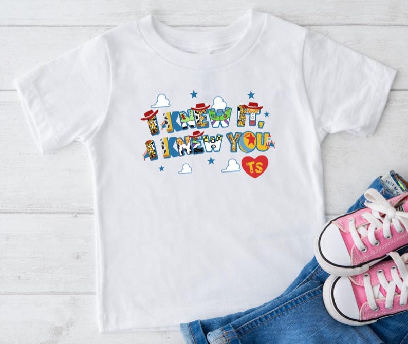 Puede incluir: Una camiseta blanca de manga corta con el texto I KNEW IT, I KNEW YOU en letras de bloque coloridas y estampadas. El dise&ntilde;o incluye peque&ntilde;os sombreros de vaquero rojos, estrellas azules, nubes blancas y un coraz&oacute;n rojo con las letras TS.