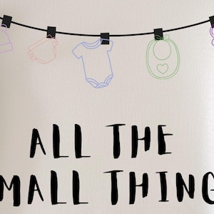 May include: A light-colored t-shirt with the phrase "ALL THE SMALL THINGS" in black lettering. Above the text, baby items like a hat, diaper, onesie, bib, and pacifier are hanging from a clothesline.