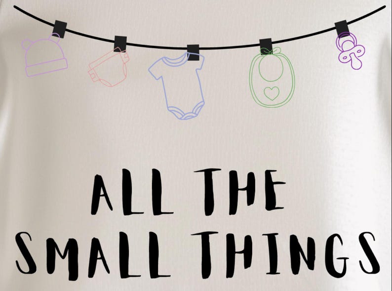 May include: A light-colored t-shirt with the phrase "ALL THE SMALL THINGS" in black lettering. Above the text, baby items like a hat, diaper, onesie, bib, and pacifier are hanging from a clothesline.