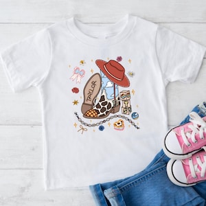 Op de afbeelding: Een wit T-shirt met korte mouwen en een grafische print van een cowboylaars met koeienprint, een rode hoed, de naam Taylor, het getal 13 en de letters BTS op blokken, omringd door kleine illustraties van sterren, bloemen en een strik.