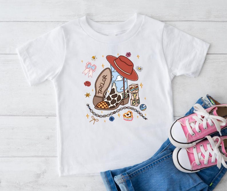 Op de afbeelding: Een wit T-shirt met korte mouwen en een grafische print van een cowboylaars met koeienprint, een rode hoed, de naam Taylor, het getal 13 en de letters BTS op blokken, omringd door kleine illustraties van sterren, bloemen en een strik.