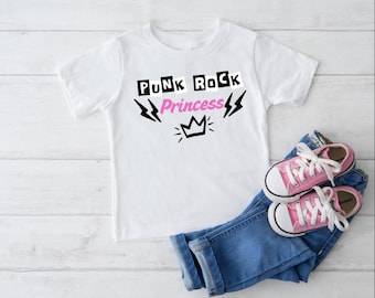 May include: A white t-shirt with the text "PUNK ROCK Princess" in black and pink, with lightning bolt and crown graphics. The shirt is paired with blue jeans and pink canvas sneakers with white laces.
