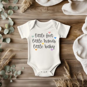 May include: White baby onesie with the text "little feet little hands little baby" in black script, accented with colorful hearts. The onesie has short sleeves and three snaps at the bottom.