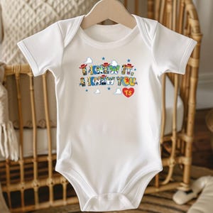 Puede incluir: Un body blanco de manga corta con un estampado gr&aacute;fico que incluye el texto I KNEW IT, A KNEW YOU y un coraz&oacute;n rojo con las letras TS. El dise&ntilde;o presenta peque&ntilde;as estrellas azules, nubes blancas y sombreros de vaquero en miniatura.