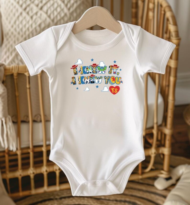 Puede incluir: Un body blanco de manga corta con un estampado gr&aacute;fico que incluye el texto I KNEW IT, A KNEW YOU y un coraz&oacute;n rojo con las letras TS. El dise&ntilde;o presenta peque&ntilde;as estrellas azules, nubes blancas y sombreros de vaquero en miniatura.