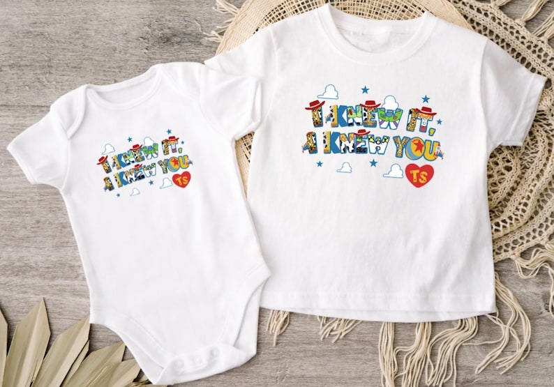 Puede incluir: Un body de beb&eacute; blanco de manga corta y una camiseta blanca de manga corta. Ambas prendas presentan un estampado gr&aacute;fico colorido con el texto "I KNEW IT, I KNEW YOU" en una fuente estilizada, acompa&ntilde;ado de peque&ntilde;as estrellas azules, nubes y un coraz&oacute;n rojo con "TS".