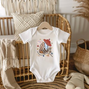 Op de afbeelding: Een wit babyrompertje met korte mouwen en een centrale afbeelding van een bruin-witte koeienprint cowboylaars met de naam Taylor, bekroond met een rode cowboyhoed, omringd door kleine illustraties van strikjes, sterretjes en bloemen.