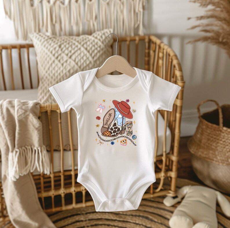 Op de afbeelding: Een wit babyrompertje met korte mouwen en een centrale afbeelding van een bruin-witte koeienprint cowboylaars met de naam Taylor, bekroond met een rode cowboyhoed, omringd door kleine illustraties van strikjes, sterretjes en bloemen.