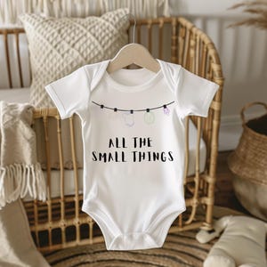 May include: White baby onesie with the text "ALL THE SMALL THINGS" in black. The onesie has a clothesline design with small illustrations of baby items. The onesie is hanging on a wooden hanger.