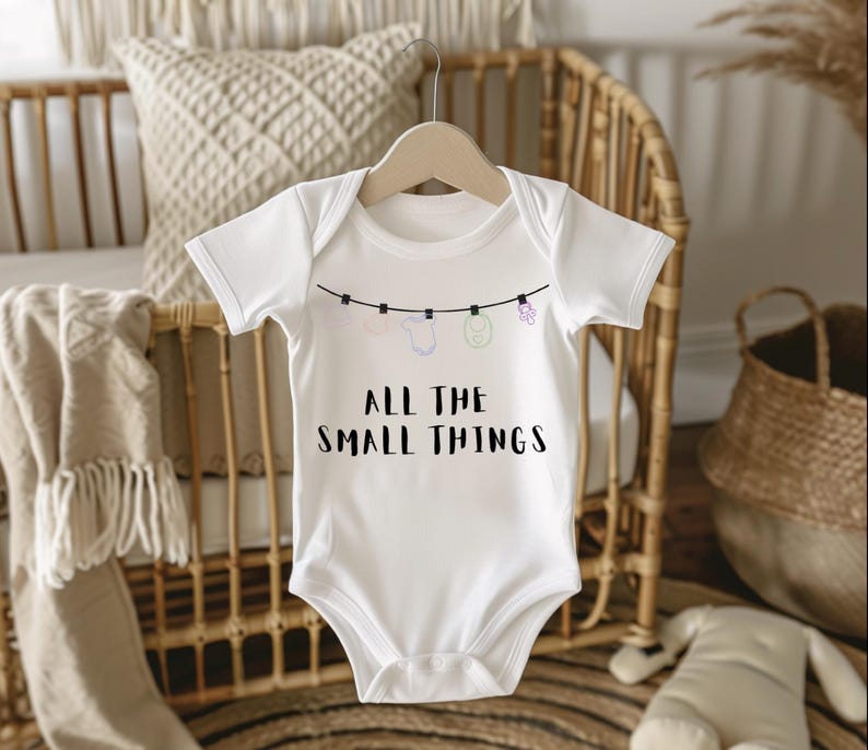 May include: White baby onesie with the text "ALL THE SMALL THINGS" in black. The onesie has a clothesline design with small illustrations of baby items. The onesie is hanging on a wooden hanger.