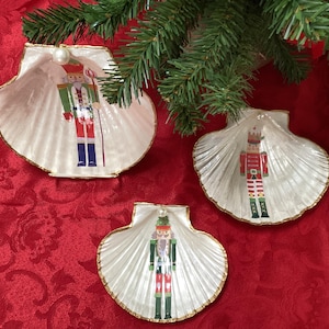 May include: Three seashell-shaped Christmas ornaments with painted nutcracker figures. The shells are white with gold trim and a pearl accent. The ornaments are arranged on a red patterned surface, with evergreen branches in the background.