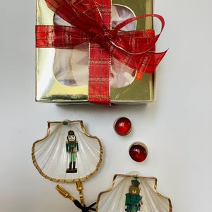 May include: A gold gift box with a red plaid ribbon and bow, alongside two white seashell-shaped ornaments with gold trim and nutcracker figures. Two red, round, jewel-like embellishments are also present.