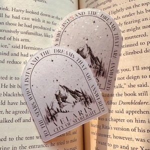 Puede incluir: Dos pegatinas arqueadas con el texto "To the stars who listen and the dreams that are answered" y "Velaris City of Starlight". Las pegatinas presentan un diseño de montaña y árboles en blanco y negro.