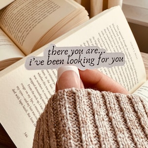 Puede incluir: Una pegatina con el texto "there you are... I've been looking for you" en cursiva. La pegatina se sostiene sobre un libro abierto con texto visible. El fondo incluye un suéter beige.