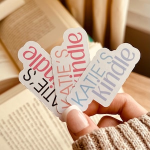 May include: Three stickers with the text "KATIE'S Kindle" in pink, light pink, and light blue. The stickers are held in front of an open book. The background is a soft, neutral colour.