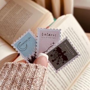 Puede incluir: Tres pegatinas decorativas con temática de libros se sostienen frente a un libro abierto. Las pegatinas son rectangulares con bordes tipo sello postal. Cada pegatina tiene un diseño y texto diferente: "Crescent City", "Velaris city of starlight" y "Basgaith way village".