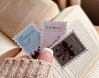 May include: Three decorative book-themed stickers are held in front of an open book. The stickers are rectangular with postage stamp-like borders. Each sticker has a different design and text: "Crescent City", "Velaris city of starlight", and "Basgaith way village".
