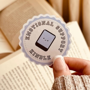 May include: A round sticker with a light blue scalloped edge. The sticker reads "EMOTIONAL SUPPORT KINDLE" in a circular pattern around a smiling Kindle graphic. The background includes open books.