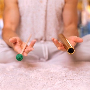 May include: A person holding a gold-colored singing bowl and a wooden mallet with a green, textured head. The bowl is cylindrical with a black interior. The mallet is made of wood and has a green, textured head. The background is blurred.