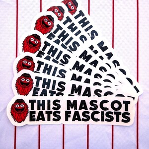 Op de afbeelding: Een verzameling witte stickers met een rood mascotte-personage en de tekst "THIS MASCOT EATS FASCISTS" in het zwart. De stickers zijn gerangschikt op een wit shirt met rode verticale strepen.