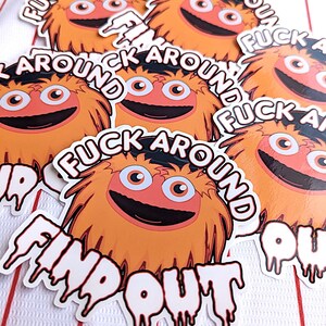 Könnte beinhalten: Eine Sammlung orangefarbener und schwarzer Aufkleber mit einer Cartoonfigur und dem Text "FUCK AROUND FIND OUT". Die Aufkleber haben einen weißen Rand und befinden sich auf einem weiß-rot gestreiften Hintergrund.