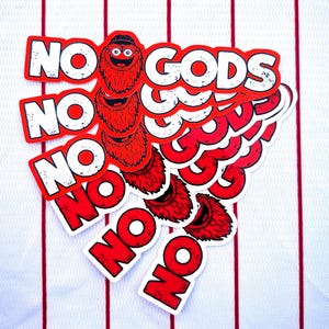 Op de afbeelding: Een verzameling rode en witte stickers met de tekst "NO GODS" en een cartoonfiguur. De stickers zijn gerangschikt op een witte stof met rode verticale strepen. Het personage is rood met een zwart gezicht en witte ogen.