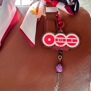 May include: A brown handbag with a red keychain featuring the text "BTS ARIRANG". The keychain has a purple bead and a silver chain. A patterned scarf is tied to the handbag's handle.