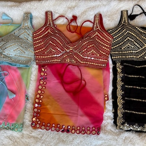 May include: Three embellished sari ensembles. The first features a light blue top with silver accents and a colorful skirt. The second has a red top and a pink and orange skirt. The third has a black top and skirt with gold accents.