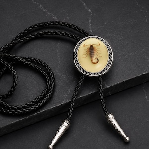 Pu&ograve; includere: Cravatta a bolo nera con un pendente circolare raffigurante uno scorpione in resina color crema, incorniciato da un bordo argentato. La cravatta ha un cordino in pelle intrecciata e punte argentate.