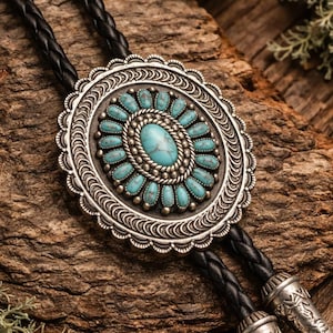 Puede incluir: Una corbata bolo plateada con una piedra turquesa central y detalles decorativos. La corbata bolo tiene un cordón de cuero trenzado negro y puntas plateadas. Sobre un fondo de madera rústica con piedras turquesas y vegetación.