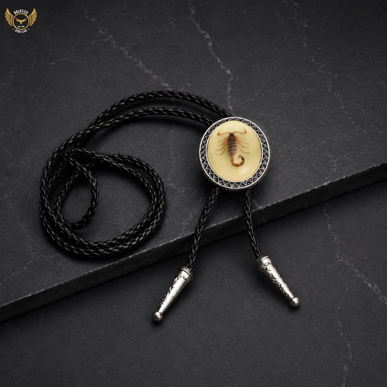 Pu&ograve; includere: Cravatta a bolo nera con un pendente circolare raffigurante uno scorpione in resina color crema, incorniciato da un bordo argentato. La cravatta ha un cordino in pelle intrecciata e punte argentate.