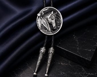 Può includere: Una cravatta a bolo argentata con un design dettagliato di testa di cavallo e piume su un ciondolo circolare. Il cordino in pelle nera ha punte argentate decorative. Lo sfondo è un tessuto blu scuro.
