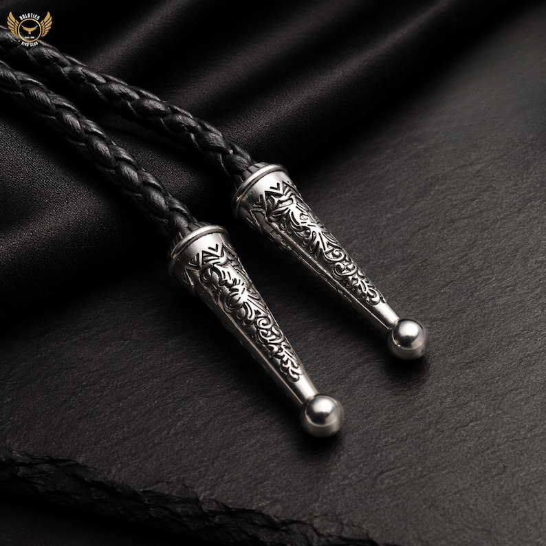 Pu&ograve; includere: Primo piano di una cravatta a bolo in pelle nera con punte decorative color argento. Le punte presentano un motivo floreale inciso e una piccola sfera rotonda all'estremit&agrave;. Il cordoncino in pelle intrecciata &egrave; visibile.