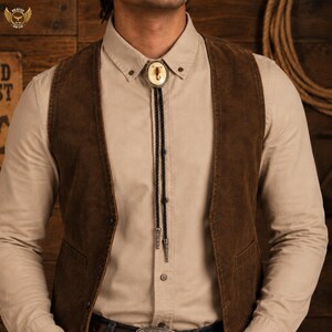Pu&ograve; includere: Una camicia beige con bottoni e un gilet in camoscio marrone sono indossati con una cravatta a bolo con un disegno di toro. La cravatta a bolo ha un cordino nero intrecciato con punte color argento. Il gilet ha una vestibilit&agrave; classica e su misura.