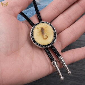 Pu&ograve; includere: Una cravatta a bolo con un ciondolo circolare raffigurante uno scorpione in resina color crema. Il ciondolo ha un bordo color argento ed &egrave; sospeso a un cordino nero intrecciato con punte decorative color argento.