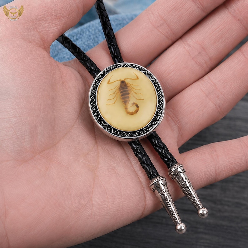 Pu&ograve; includere: Una cravatta a bolo con un ciondolo circolare raffigurante uno scorpione in resina color crema. Il ciondolo ha un bordo color argento ed &egrave; sospeso a un cordino nero intrecciato con punte decorative color argento.