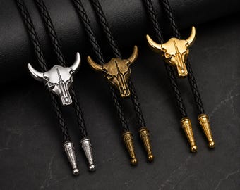 Può includere: Tre cravatte a bolo con ciondoli a forma di teschio di toro argentati, bronzo e dorati. Ogni cravatta ha un cordino nero intrecciato e puntali metallici decorativi. Presentate su una superficie scura.