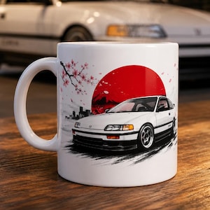 May include: White ceramic mug featuring a Japanese-inspired design with a red sun, cherry blossoms, and a white sports car. The mug has a curved handle and a glossy finish, perfect for coffee or tea.