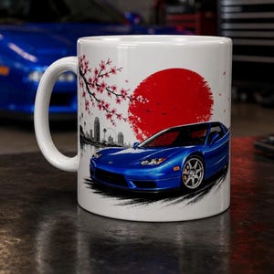 May include: White ceramic mug featuring a blue sports car design with a red sun, cherry blossoms, and a cityscape. The mug has a curved handle and is suitable for hot or cold drinks.
