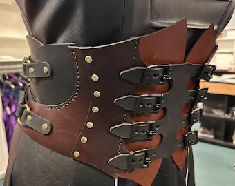 May include: A brown and black leather corset with multiple buckles and rivets. The corset has a layered design with a dark brown center panel and black side panels. The buckles are black and the rivets are gold.