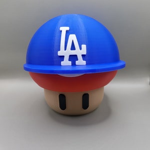 May include: A 3D printed mushroom figurine with a blue baseball cap with the LA logo in white. The mushroom is red and brown.