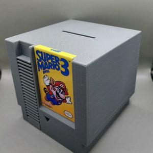 May include: A gray 3D printed piggy bank shaped like a Nintendo Entertainment System (NES) game console. The bank has a slot on the top for inserting coins. A Super Mario Bros. 3 game cartridge is inserted into the front of the bank.